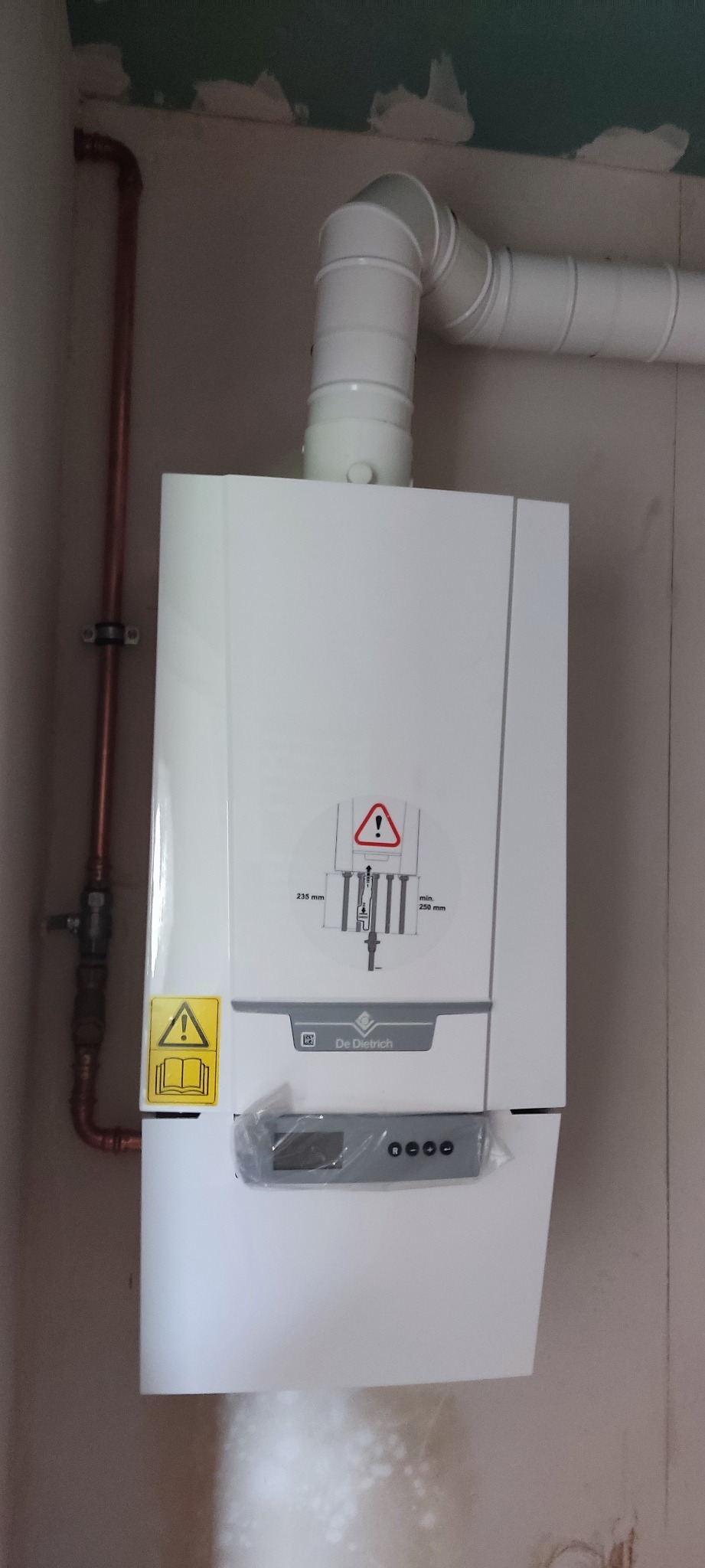 Nowy, biały piec gazowy marki De Dietrich z podłączonymi rurami miedzianymi i białym kominem, jeszcze w folii ochronnej na wyświetlaczu, zamontowany na nierównej ścianie.