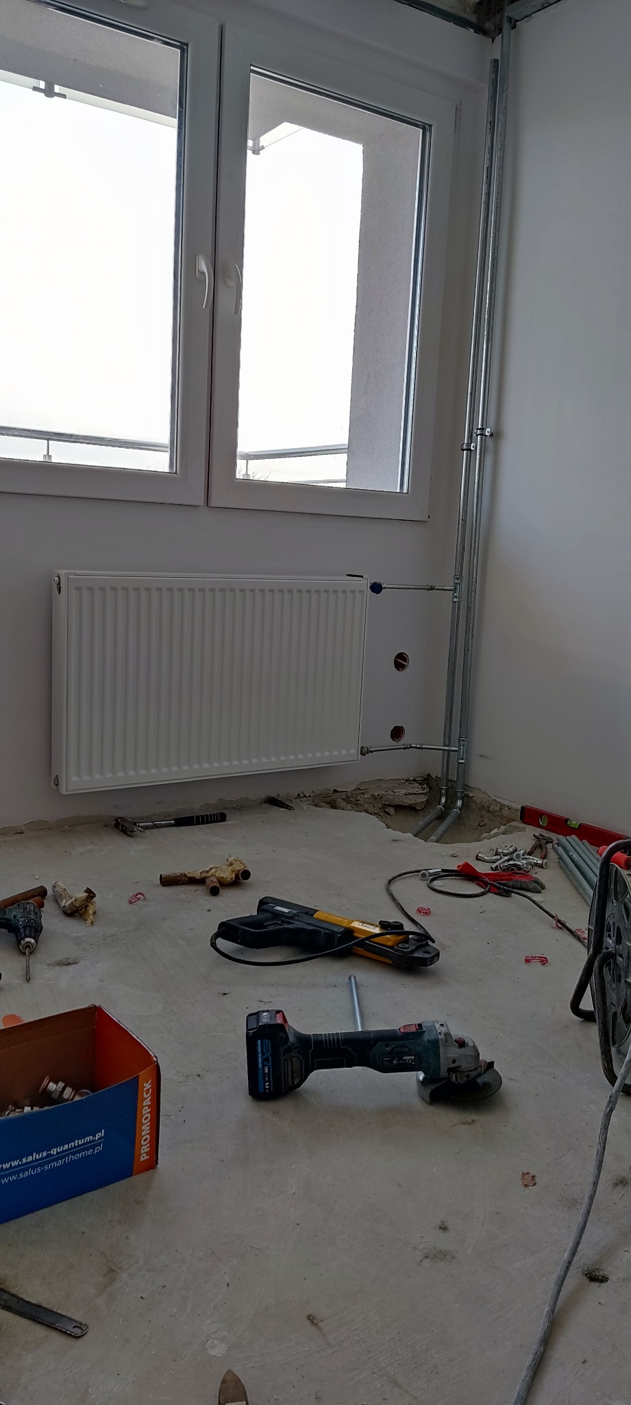 Wnętrze w trakcie montażu instalacji grzewczej: biały grzejnik pod oknem, rury instalacji wyprowadzone ze ściany, narzędzia i akcesoria hydrauliczne rozłożone na surowej podłodze.