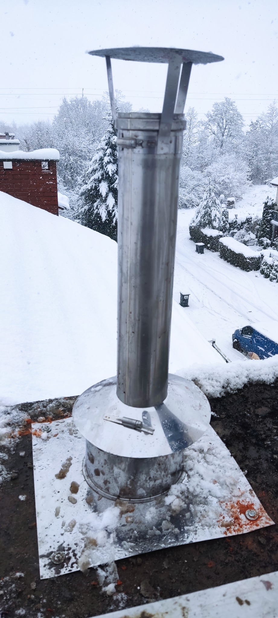Nowy, błyszczący, metalowy komin na dachu pokrytym śniegiem podczas opadów śniegu. W tle ośnieżone drzewa i budynki.