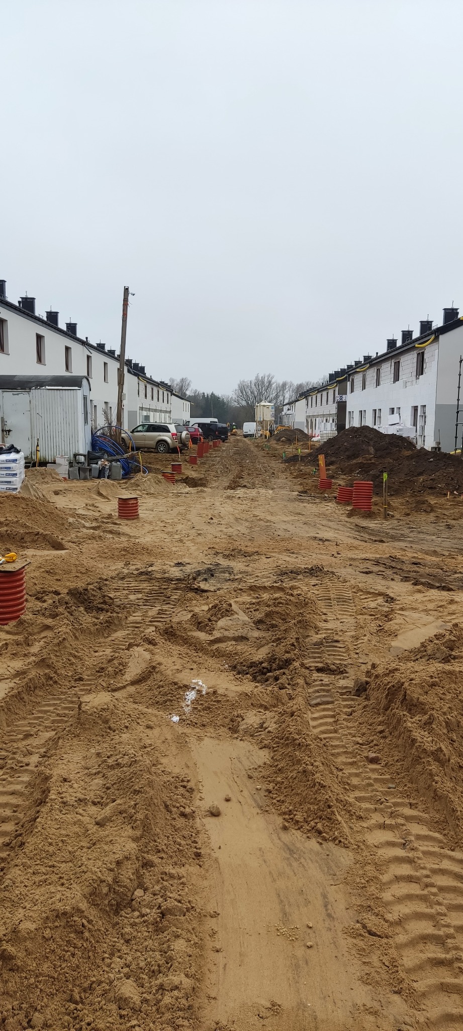 Ulica w trakcie budowy osiedla domów szeregowych, widoczne wykopane rury kanalizacyjne, piasek i maszyny budowlane.