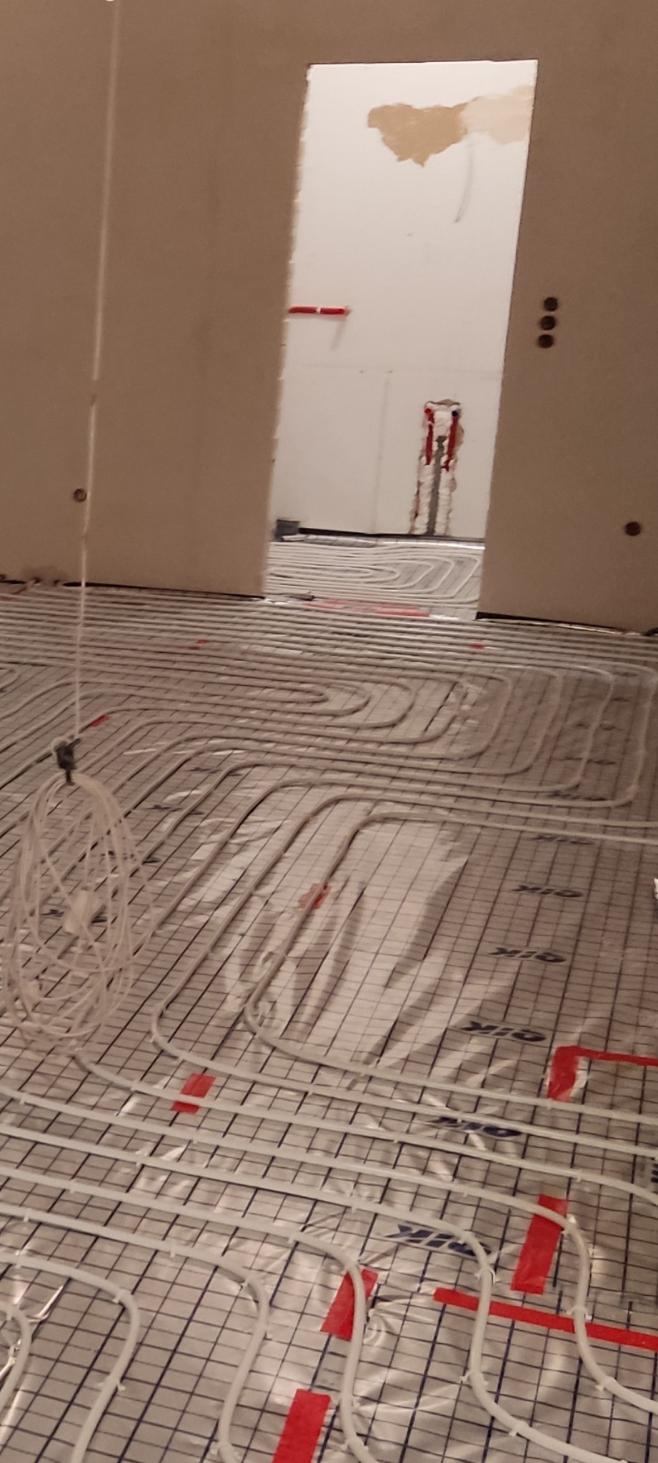 Białe rury systemu ogrzewania podłogowego ułożone na folii z siatką w surowym wnętrzu, widoczne czerwone taśmy mocujące i przewody zwisające z sufitu, w tle otwór drzwiowy z instalacją.