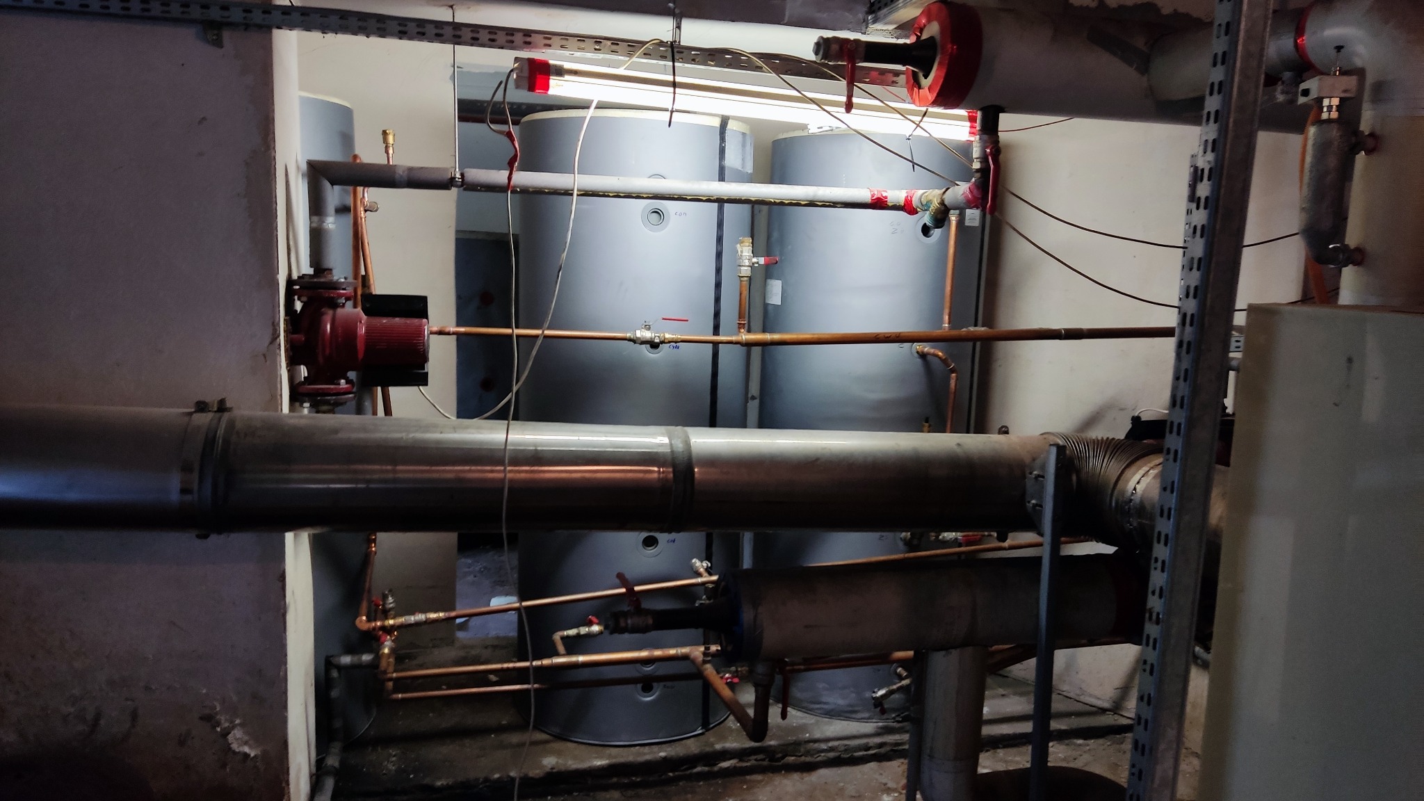 Kotłownia z dwoma szarymi zbiornikami akumulacyjnymi, rurami miedzianymi i stalowymi, pompą obiegową z czerwonym korpusem oraz oświetleniem jarzeniowym.