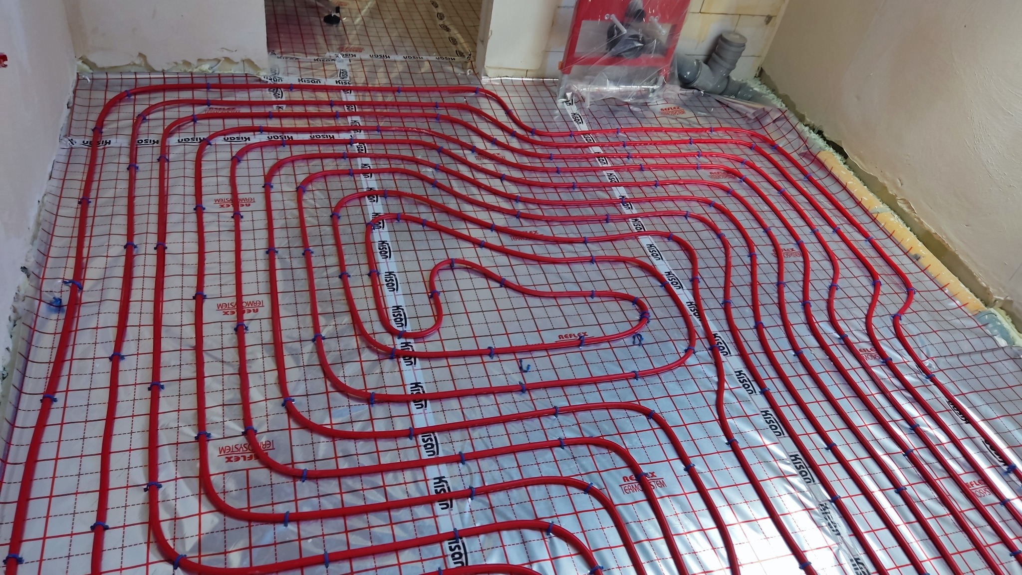 Czerwone rury grzewcze ułożone w pętle na folii z siatką, przygotowane do zalania w posadzce ogrzewania podłogowego, widoczne niebieskie mocowania.