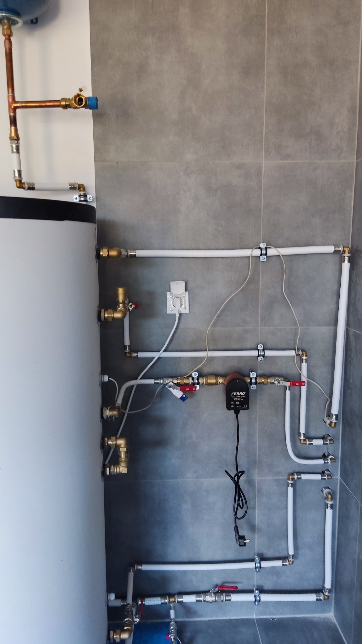Kotłownia z białym zasobnikiem ciepłej wody, licznymi miedzianymi i plastikowymi rurami, zaworami, pompą obiegową FERRO oraz szarą, płytkową ścianą w tle.