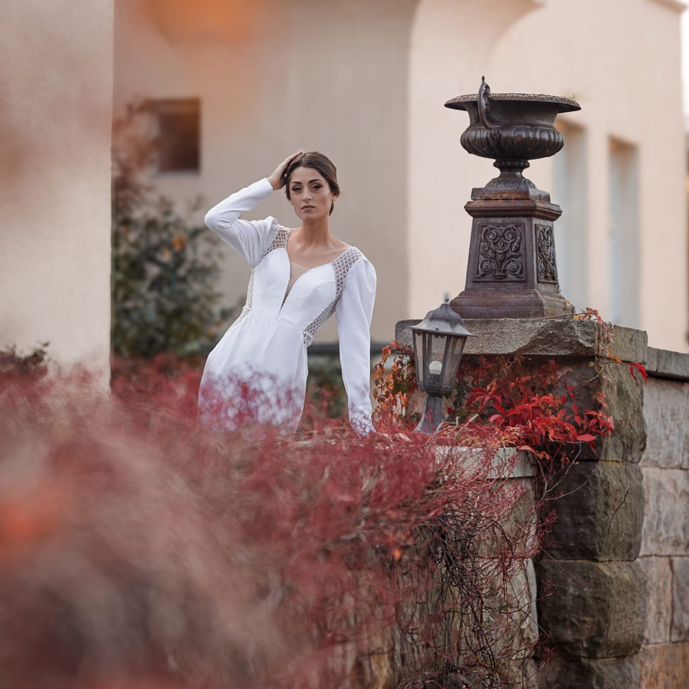 Modelka w białej sukni z długimi rękawami i koronkowymi wstawkami, pozująca na tle kamiennego murku z dekoracyjną urną i lampionem, otoczona czerwonymi krzewami.