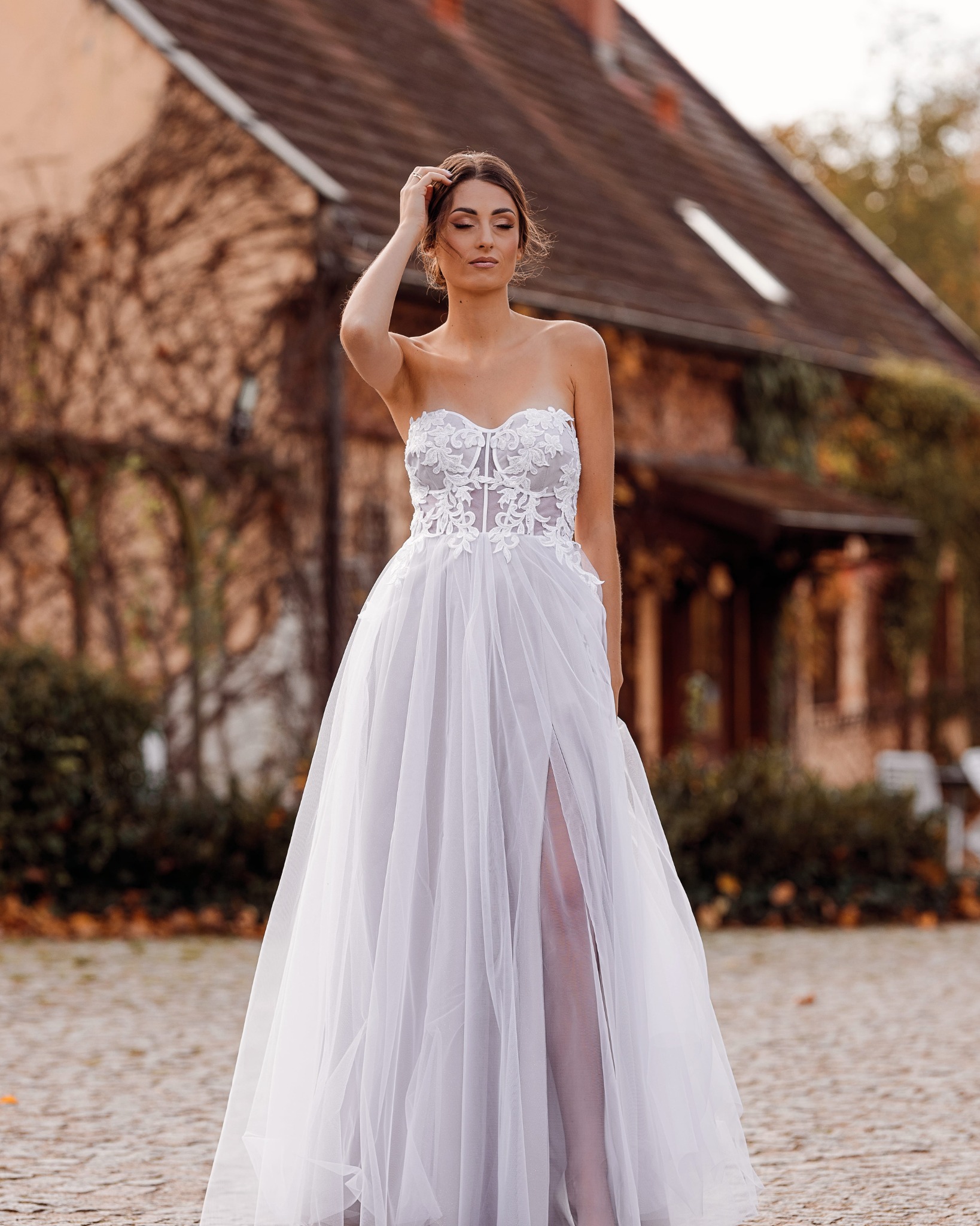 Panna młoda w zwiewnej białej sukni z koronkowym gorsetem i rozcięciem na tle ceglanego budynku porośniętego bluszczem.