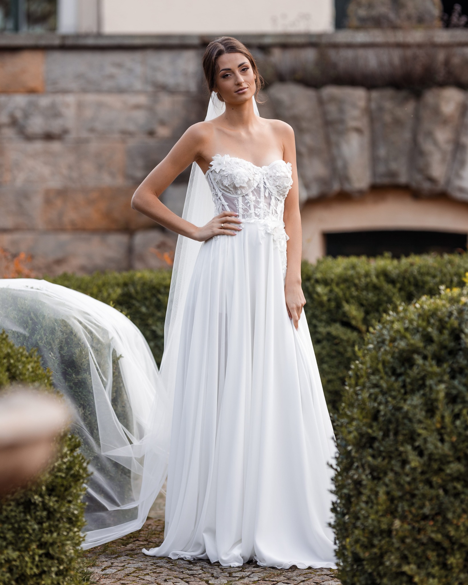 Portret panny młodej w białej sukni bez ramiączek, z koronkowym gorsetem i długim welonem, pozującej w ogrodzie na tle kamiennej ściany.