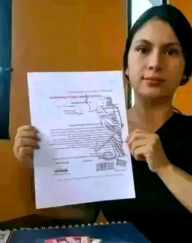 Kobieta trzyma w rękach dokument z rysunkiem Temidy i tekstem prawnym, prezentowany na tle żółtej ściany.