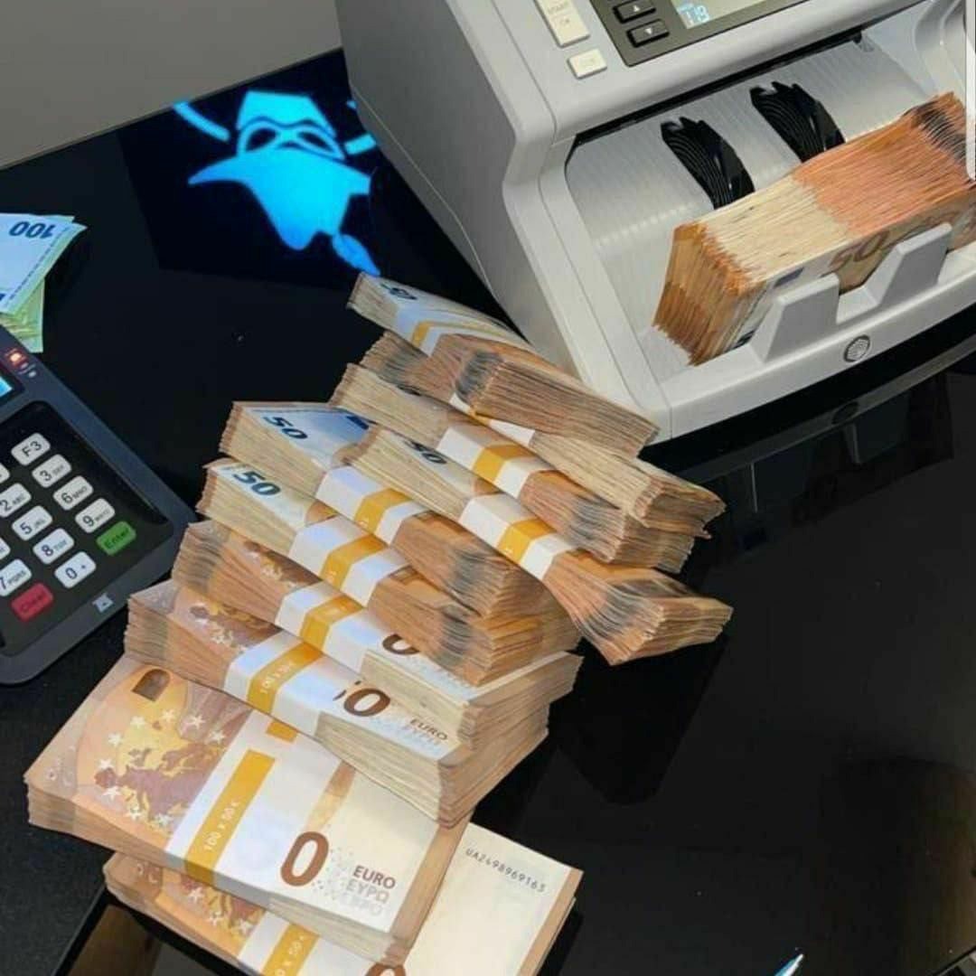 Stosy banknotów euro o nominałach 50 i 100 euro, spięte banderolami, leżące na błyszczącej, czarnej powierzchni obok licznika banknotów i terminala płatniczego. Na powierzchni widoczne odbicie...