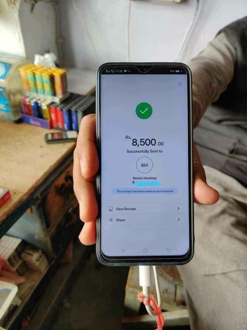 Ekran smartfona z potwierdzeniem pomyślnego przelewu kwoty 8500 Rs. do Benish Mushtaq, widoczny kabel ładowarki. W tle półka z różnymi przedmiotami.