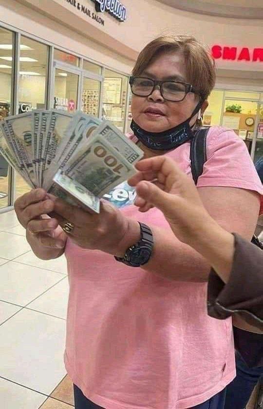 Kobieta w różowej koszulce i okularach trzyma wachlarz banknotów studolarowych, druga osoba wyciąga rękę po pieniądze w centrum handlowym.