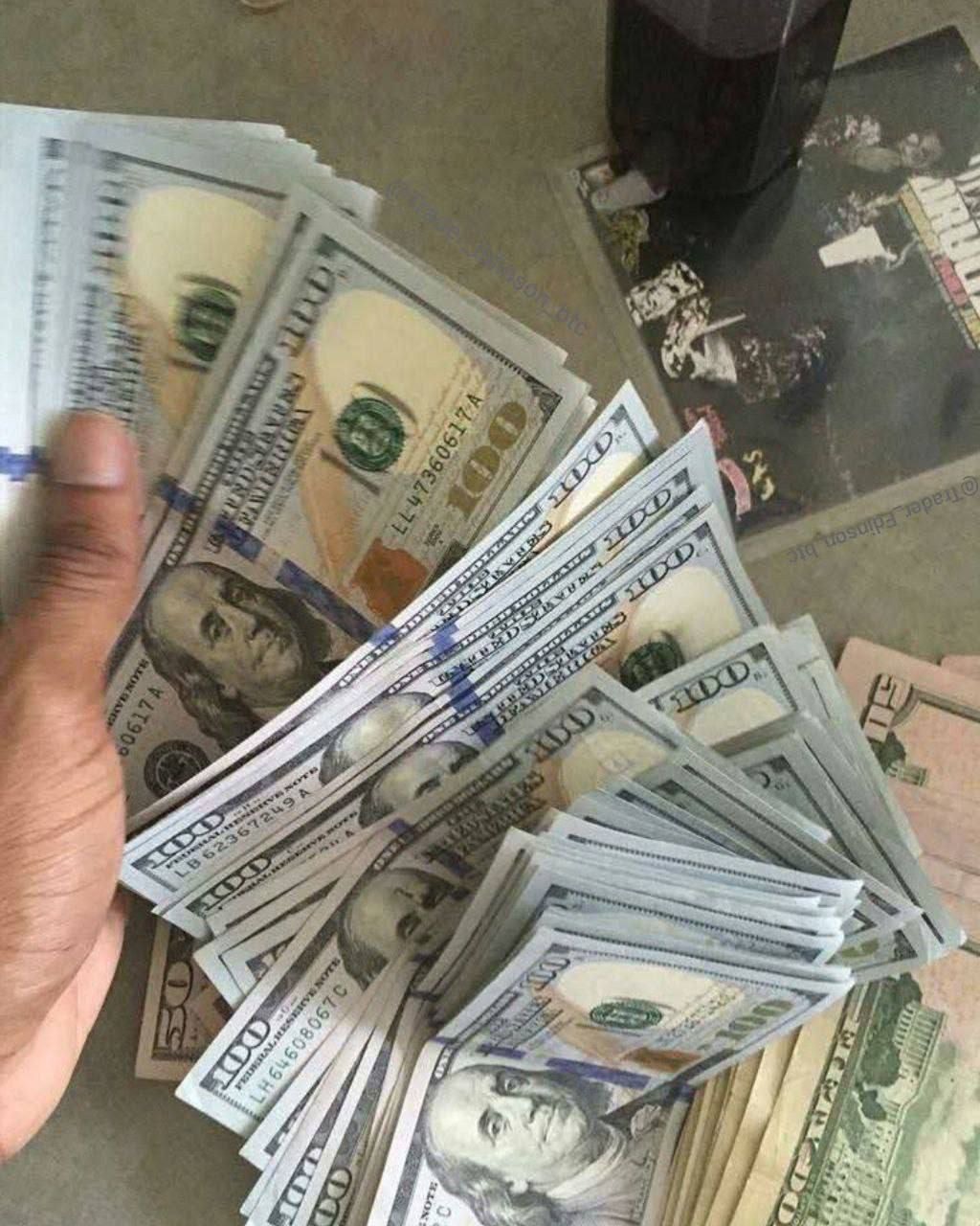 Stos banknotów studolarowych ułożonych na stole obok szklanki z ciemnym napojem i okładki płyty winylowej, częściowo zasłonięty dłonią.