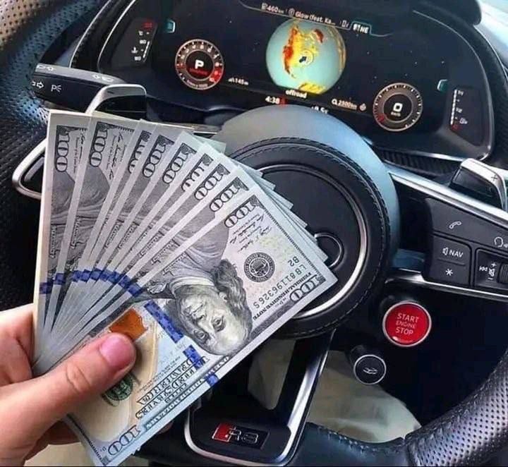 Stos banknotów studolarowych trzymanych w dłoni na tle kierownicy i deski rozdzielczej samochodu Audi R8 z włączonym ekranem nawigacji.