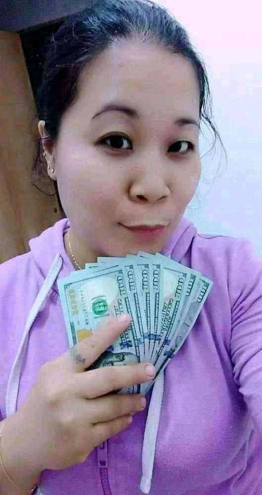 Uśmiechnięta kobieta w fioletowej bluzie trzymająca wachlarz banknotów studolarowych, z widocznym tatuażem na palcu wskazującym.