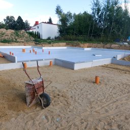 Zakład Budowlany - Przygotowana płyta fundamentowa z izolacją termiczną, widoczne pionowe rury instalacyjne i taczka na piasku. W tle budynek w stanie surowym otwartym i drzewa.