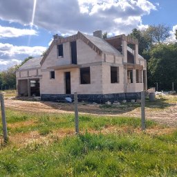 Zakład Budowlany - Surowy stan domu w trakcie budowy z białych bloczków, widoczne niezabezpieczone otwory okienne i drzwiowe, częściowo zadaszony, na tle zieleni i błękitnego nieba z chmurami.