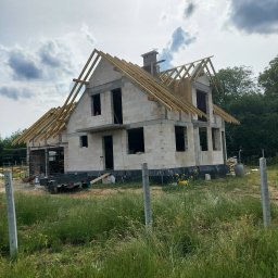 Zakład Budowlany - Surowy stan dwukondygnacyjnego domu z białych bloczków, z widoczną konstrukcją dachu i kominem, na tle pochmurnego nieba, z przyczepką i narzędziami budowlanymi w pobliżu.