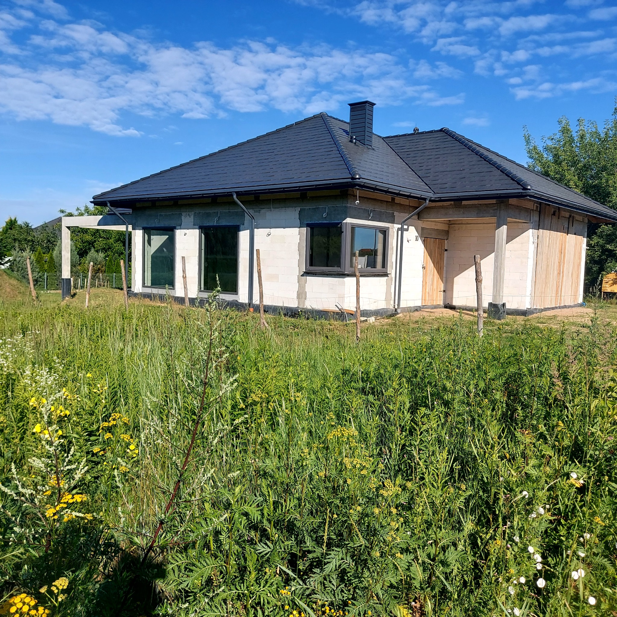 Surowy stan domu z białego bloczka z czarnym dachem, widoczne drewniane panele i wysoka trawa na pierwszym planie.