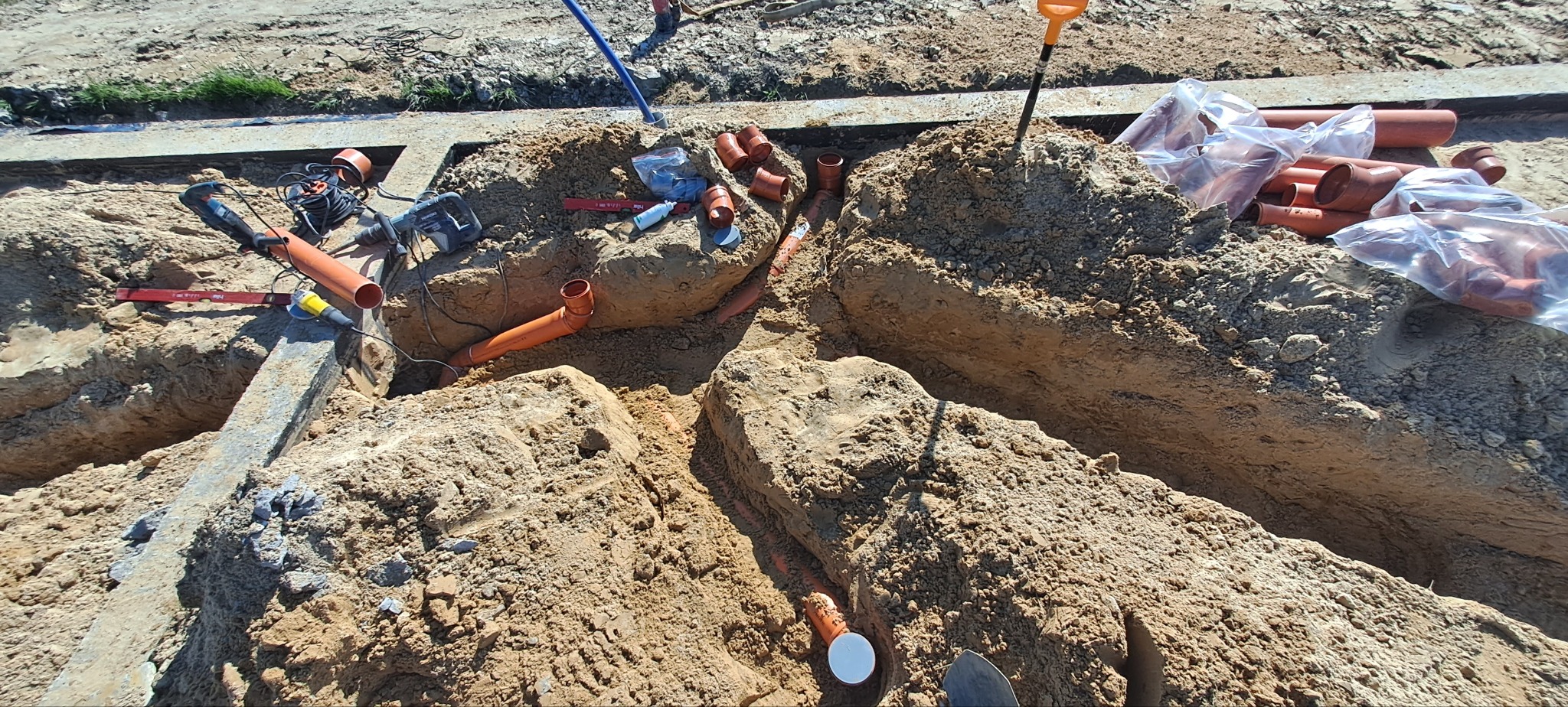 Wykop z pomarańczowymi rurami kanalizacyjnymi, narzędziami i akcesoriami hydraulicznymi w trakcie instalacji.