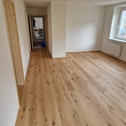 WULK-BUD - Jasne panele podłogowe z widocznymi słojami w nowym, pustym pokoju z białymi ścianami i drewnianymi futrynami drzwi, widziane z perspektywy szerokokątnej.