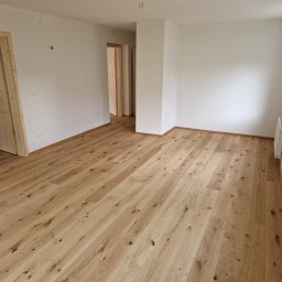 WULK-BUD - Jasne, drewniane panele podłogowe z widocznymi słojami w pustym, odnowionym pokoju z białymi ścianami i dwiema parami gniazdek elektrycznych. Widoczne drzwi w tle.