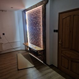 WULK-BUD - Wykończone wnętrze z dekoracyjną ścianą z cegły, podświetloną taśmą LED, drewnianym blatem i panelami podłogowymi. Widoczny fragment drzwi i przygotowane panele do montażu.