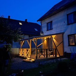 WULK-BUD - Drewniana, oświetlona weranda z balustradą w kształcie litery X, przylegająca do domu z oknami. Wieczorny kadr z ogrodem i widocznym fragmentem sąsiedniego budynku.