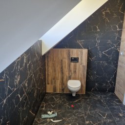 WULK-BUD - Łazienka na poddaszu z czarnymi płytkami imitującymi marmur i drewnianą zabudową toalety. Ukończony remont z widocznymi narzędziami na podłodze.