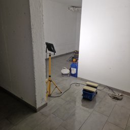 WULK-BUD - Przygotowania do malowania: lampa na statywie, kuweta malarska i farby w pomieszczeniu z szarą, ceramiczną podłogą. Widoczny fragment pomalowanej ściany.