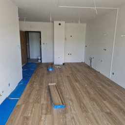 WULK-BUD - Wnętrze w trakcie układania paneli podłogowych. Część podłogi pokryta panelami o wzorze drewna, widoczne rozłożone opakowania paneli i niebieska folia izolacyjna.