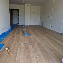 WULK-BUD - Układanie paneli podłogowych w jasnym wnętrzu. Widoczne narzędzia: młotek, kątownik, częściowo ułożone panele i podkład. Ślady robocze na powierzchni.