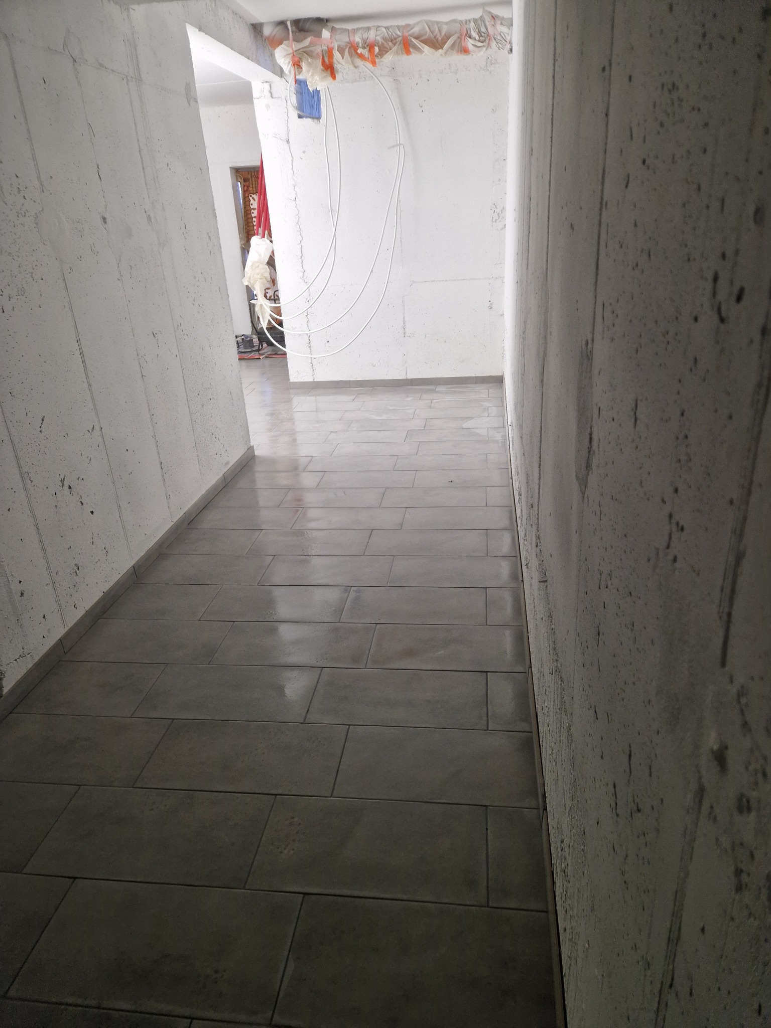 Ułożona, szara podłoga z płytek w surowym wnętrzu. Widoczne betonowe ściany i wystające kable. Perspektywa z dołu podkreśla długość korytarza.