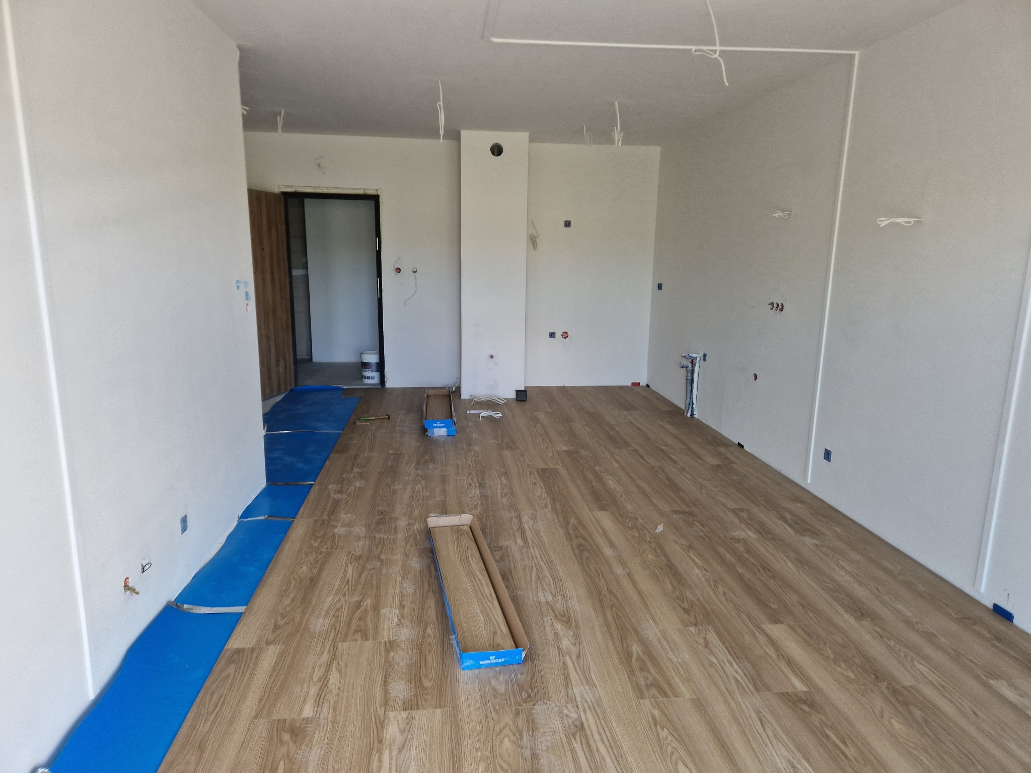 Wnętrze w trakcie układania paneli podłogowych. Część podłogi pokryta panelami o wzorze drewna, widoczne rozłożone opakowania paneli i niebieska folia izolacyjna.