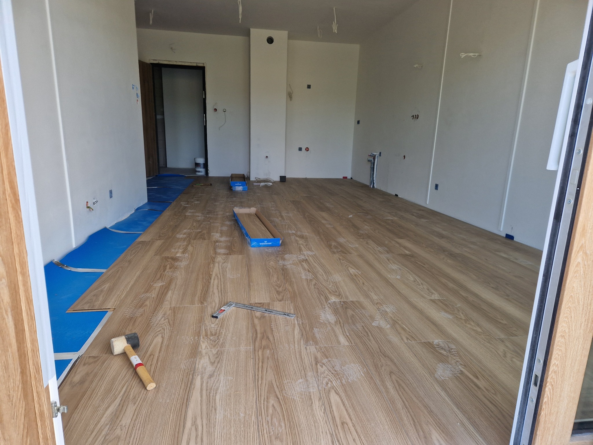 Układanie paneli podłogowych w jasnym wnętrzu. Widoczne narzędzia: młotek, kątownik, częściowo ułożone panele i podkład. Ślady robocze na powierzchni.