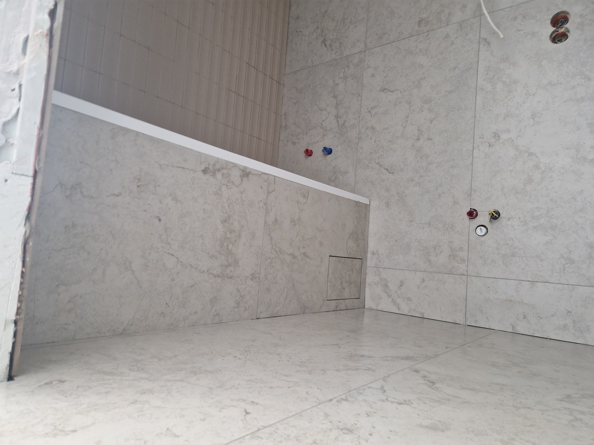 Wykończona łazienka z jasnymi, marmurkowymi płytkami na ścianach i podłodze. Widoczne przyłącza wodne i fragment wanny z białą listwą wykończeniową.