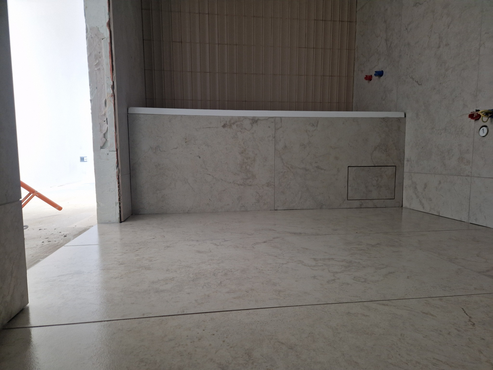 Wykończona łazienka z beżowymi płytkami podłogowymi i ściennymi. Widoczne fragmenty instalacji wodnej i fragment wanny. Częściowo wykończone ściany.