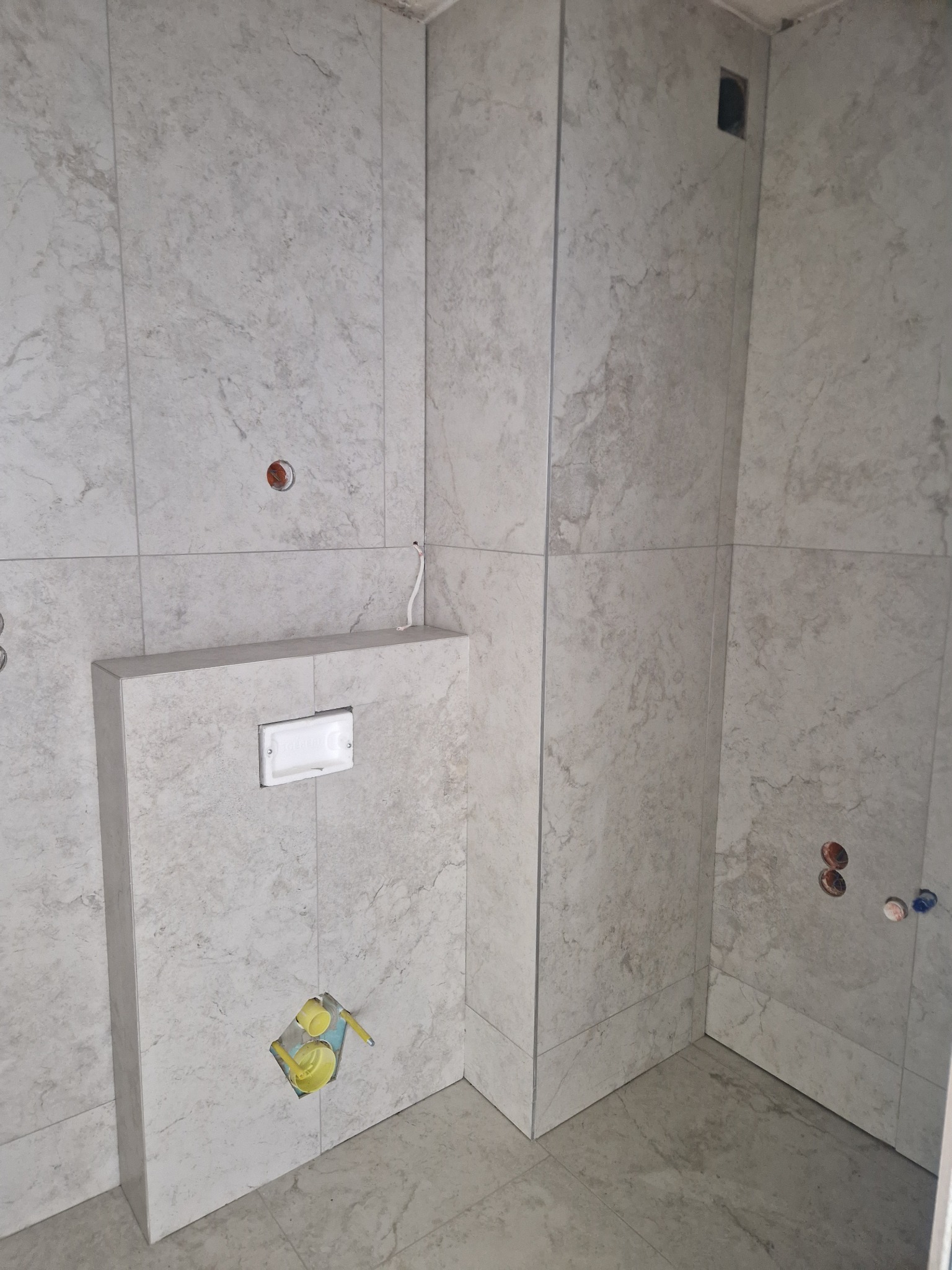 Wykończona łazienka z marmurowymi płytkami, widoczne przyłącza wodne i zabudowa stelaża WC. Minimalistyczny design, jasne barwy, surowy styl.