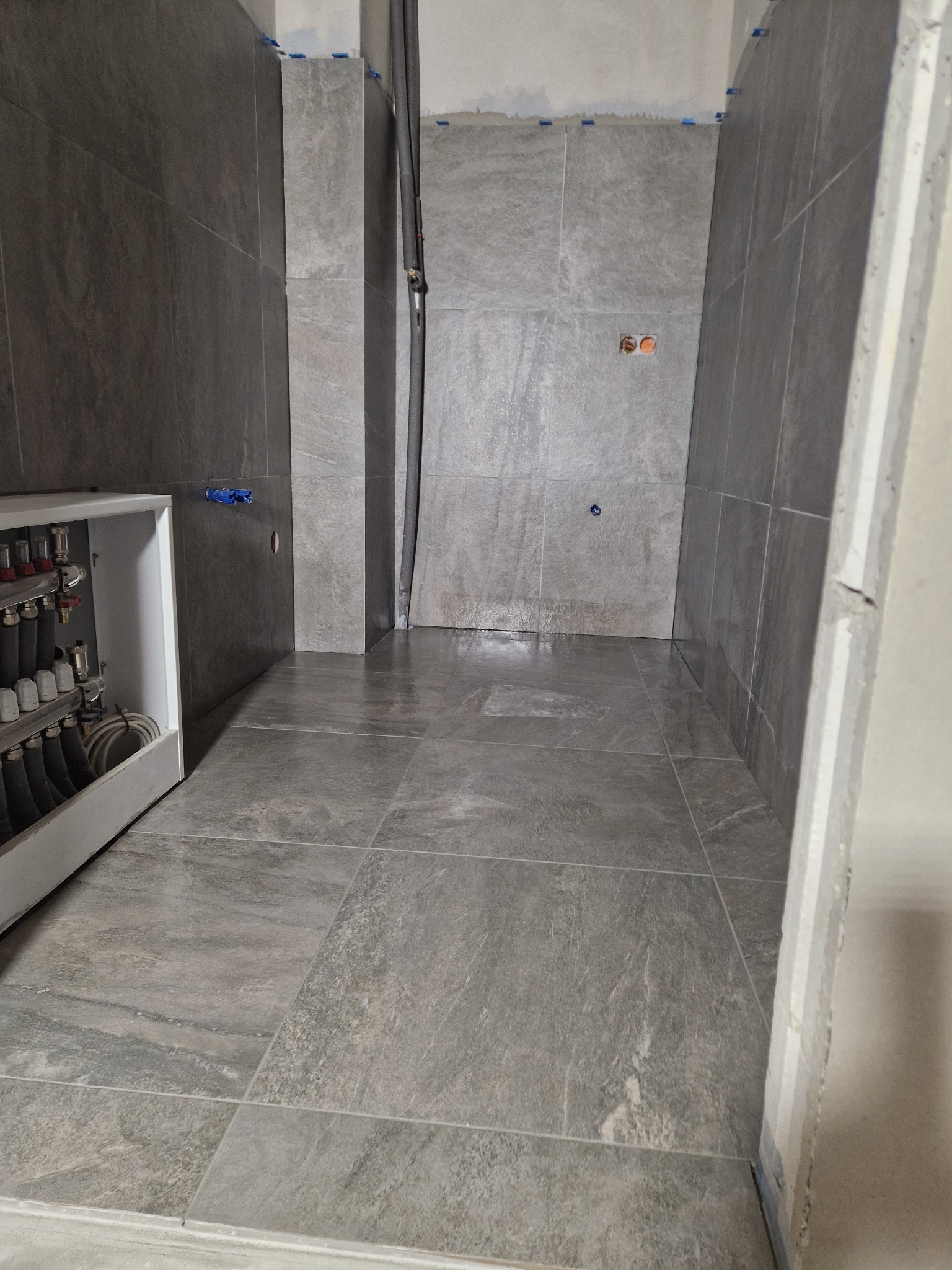 Wykończona łazienka z szarymi płytkami imitującymi kamień na ścianach i podłodze. Widoczna instalacja grzewcza i rury. Minimalistyczny design, surowe wykończenie.