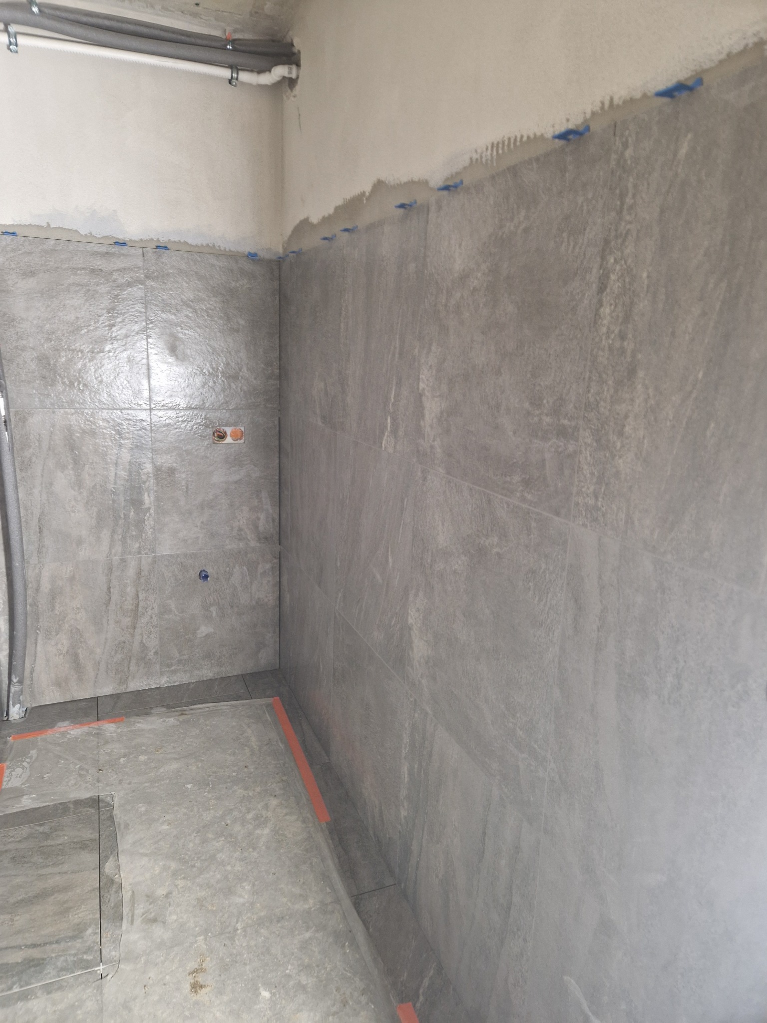 Ściana łazienki w trakcie wykańczania, pokryta szarymi płytkami imitującymi kamień. Widoczne niebieskie kliny dystansowe i instalacja elektryczna. Podłoga surowa, przygotowana do dalszych prac.