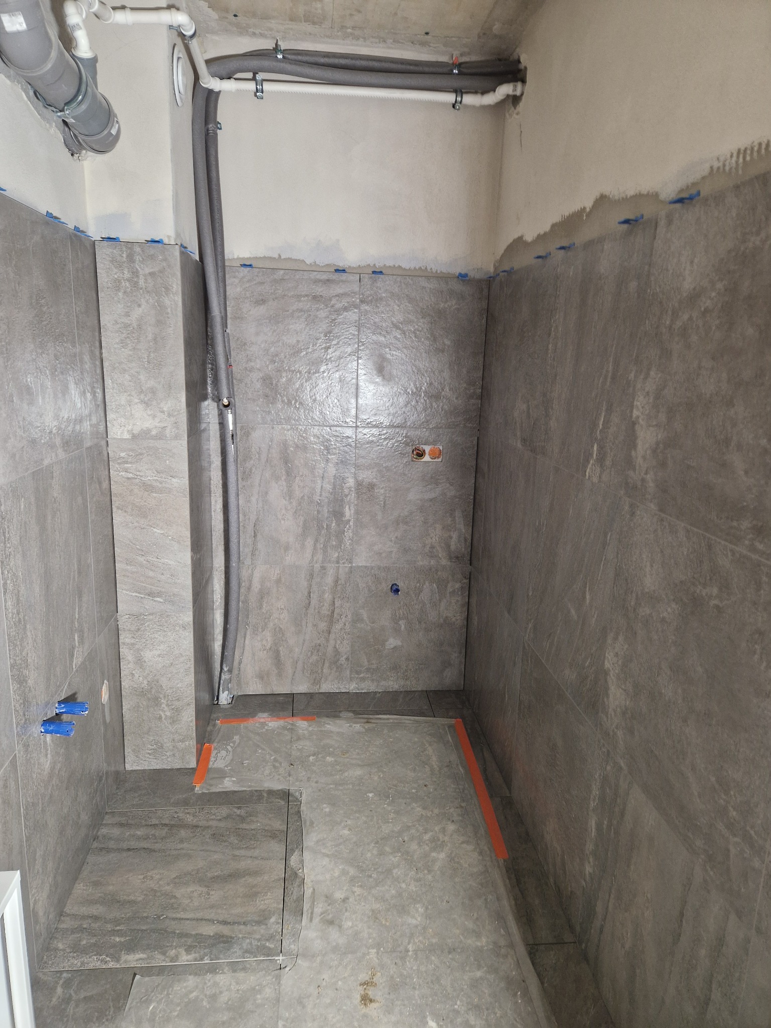 Szare płytki imitujące kamień na ścianach łazienki w trakcie remontu. Widoczne rury, niebieskie krzyżyki dystansowe i pomarańczowa taśma wyznaczająca obszar.