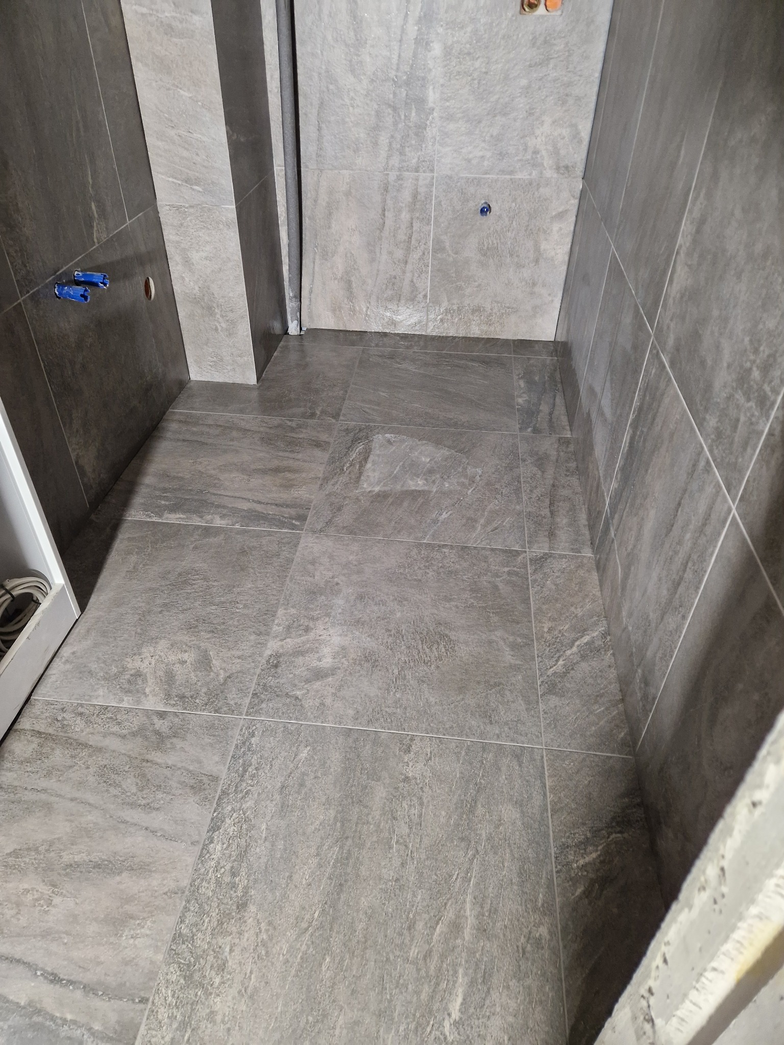 Wykończona łazienka z szarymi, matowymi płytkami imitującymi kamień na ścianach i podłodze. Widoczne fugi i elementy instalacji. Minimalistyczny design.