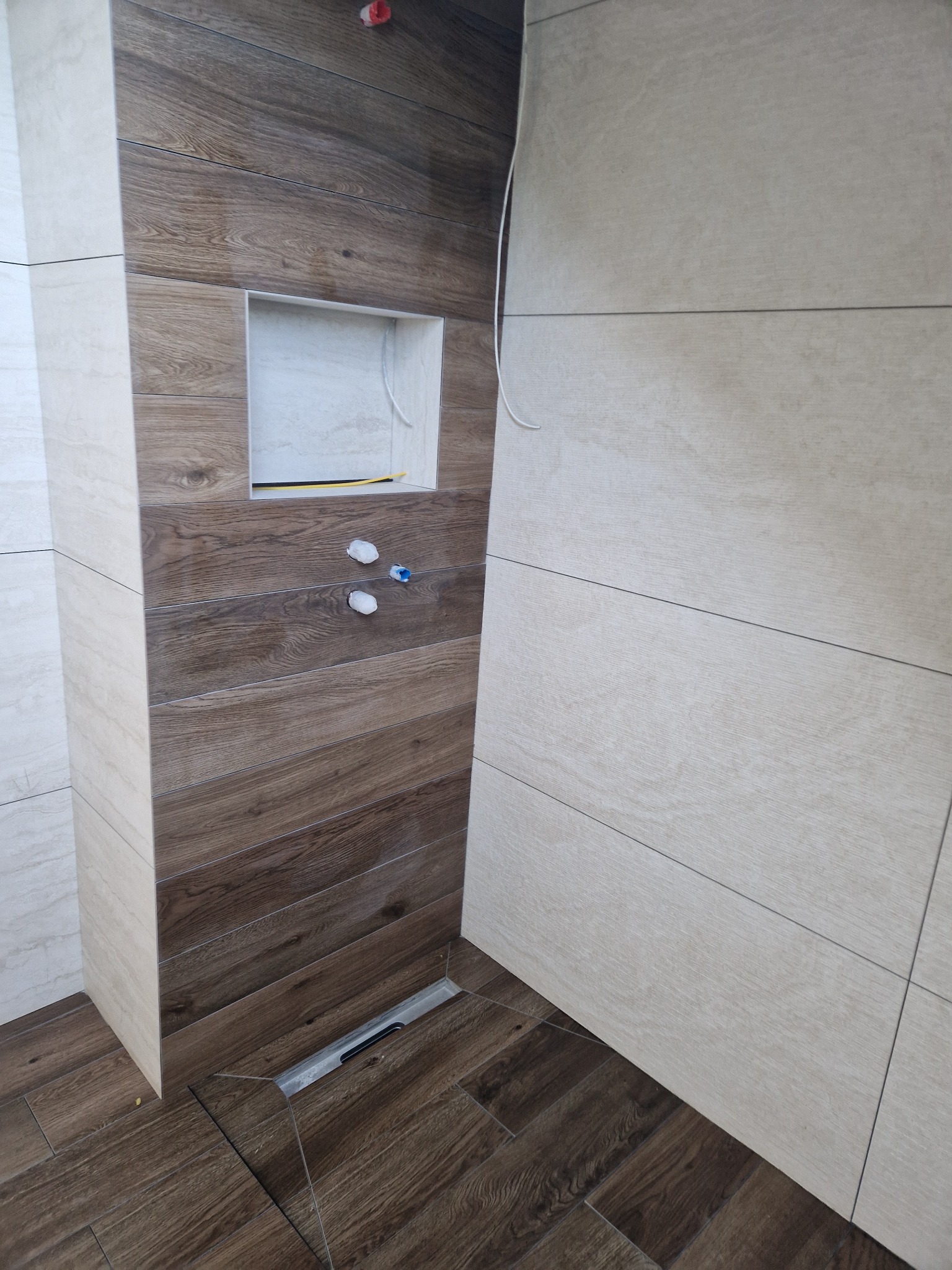 Wykończona łazienka z drewnopodobnymi płytkami na ścianach i podłodze, widoczna wnęka w ścianie i odpływ liniowy w podłodze.