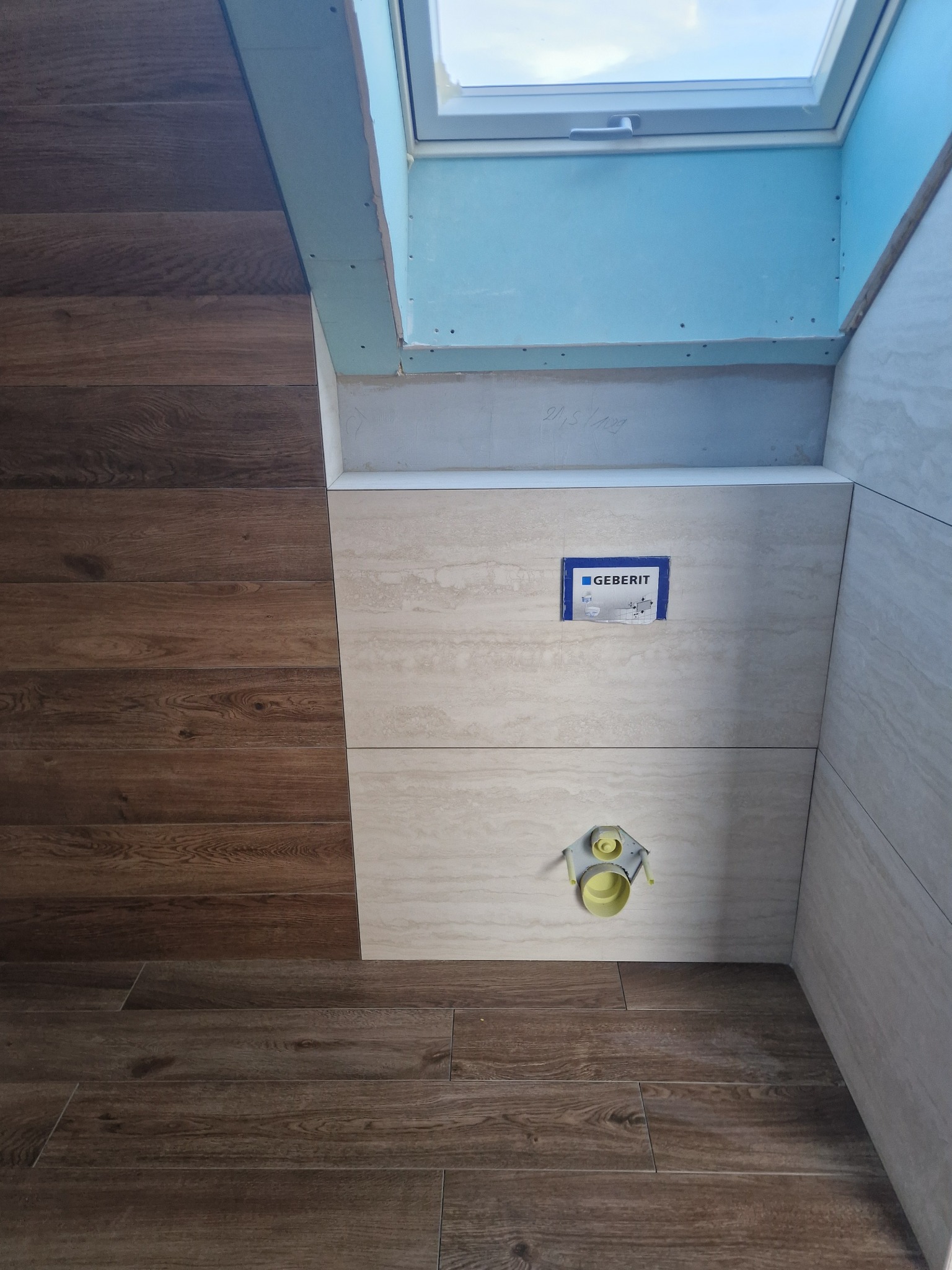 Łazienka w trakcie wykończenia: brązowe panele podłogowe, beżowe płytki ścienne, stelaż podtynkowy Geberit i okno dachowe. Widoczne elementy instalacji.