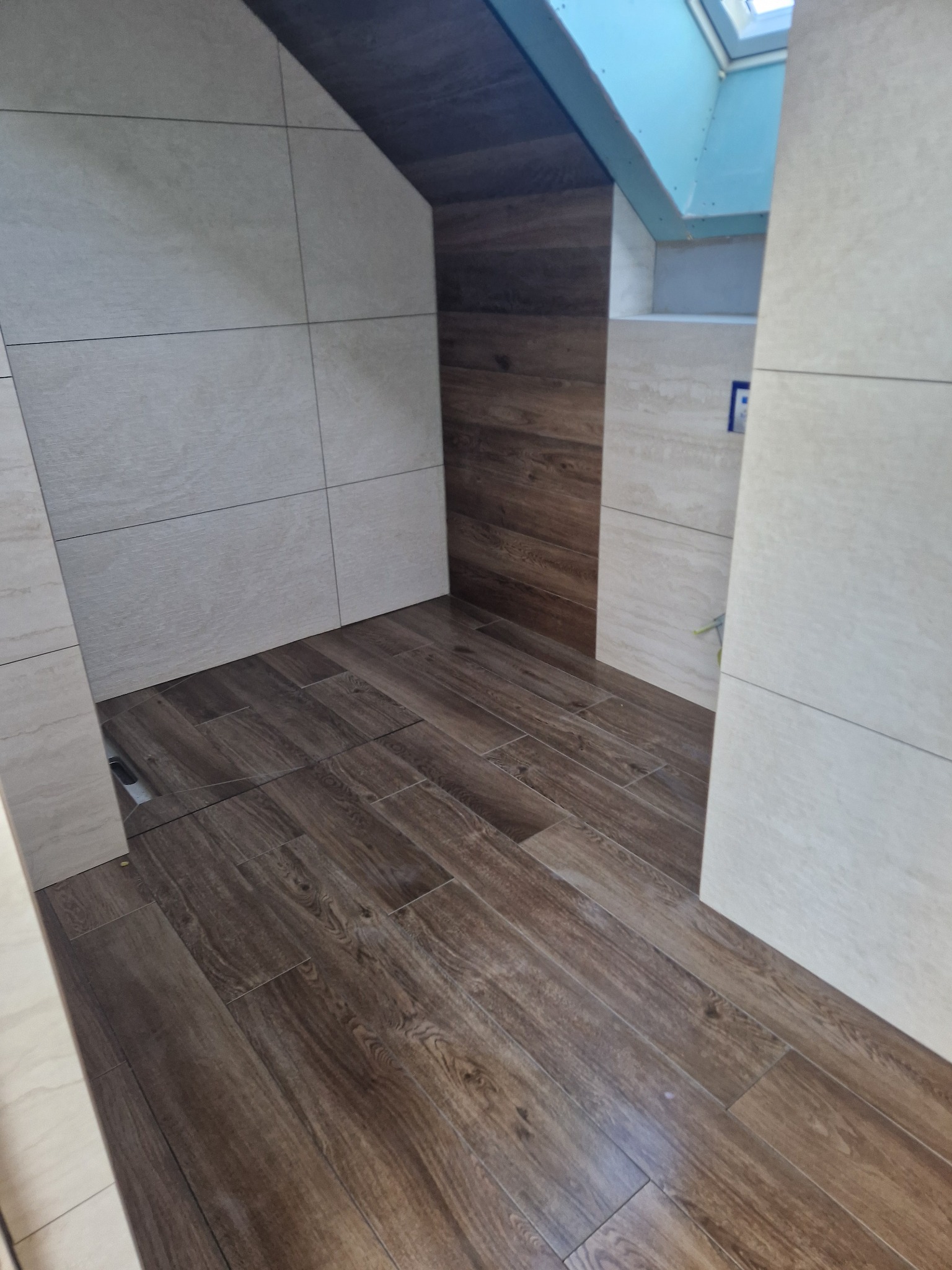 Wykończona łazienka na poddaszu z drewnopodobną podłogą i jasnymi płytkami ściennymi. Widoczny fragment okna dachowego i skośny sufit.