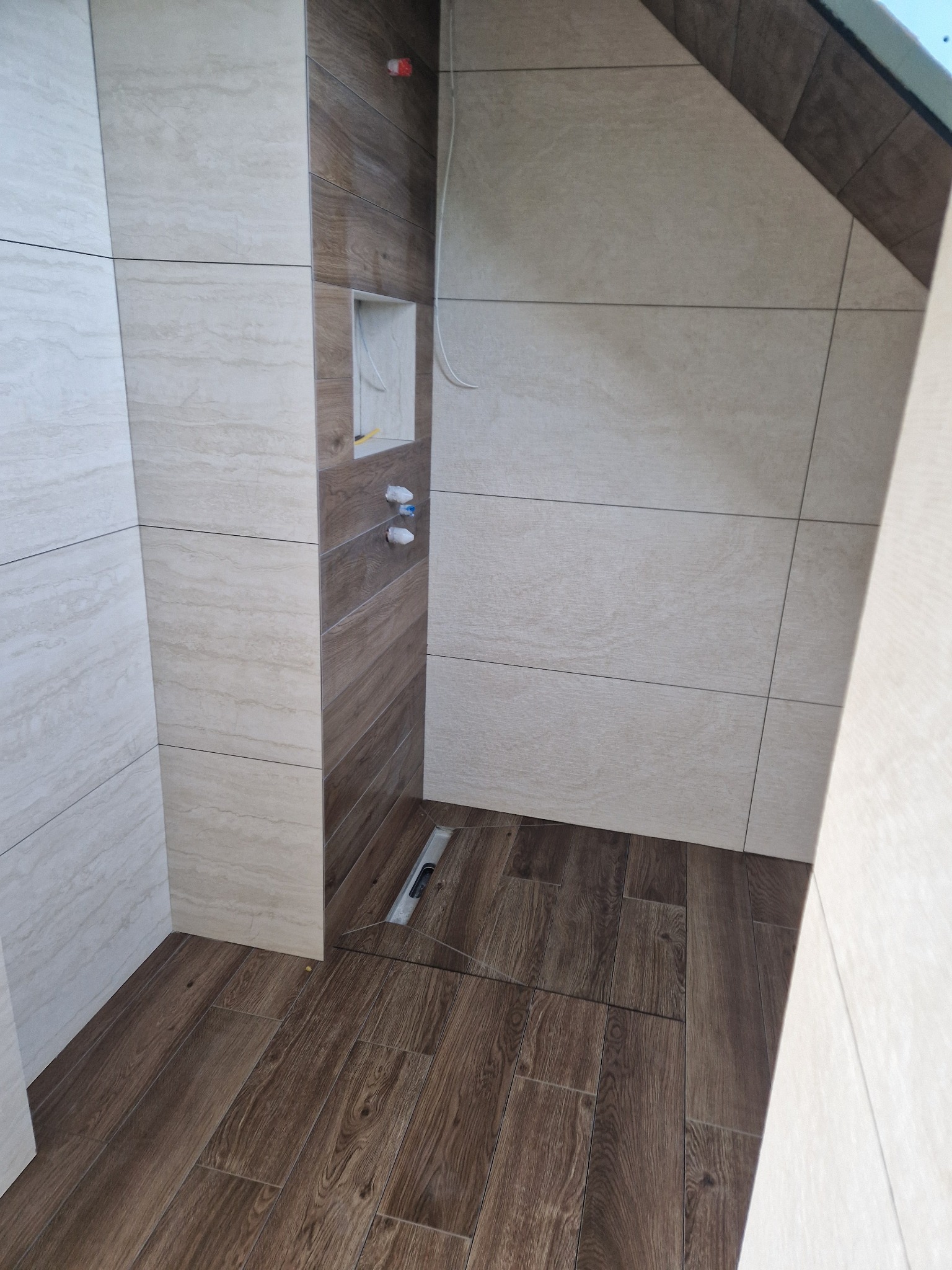 Wykończona łazienka z brązowymi panelami podłogowymi imitującymi drewno i jasnymi płytkami ściennymi z delikatnym wzorem. Widoczny odpływ liniowy i wnęka w ścianie.