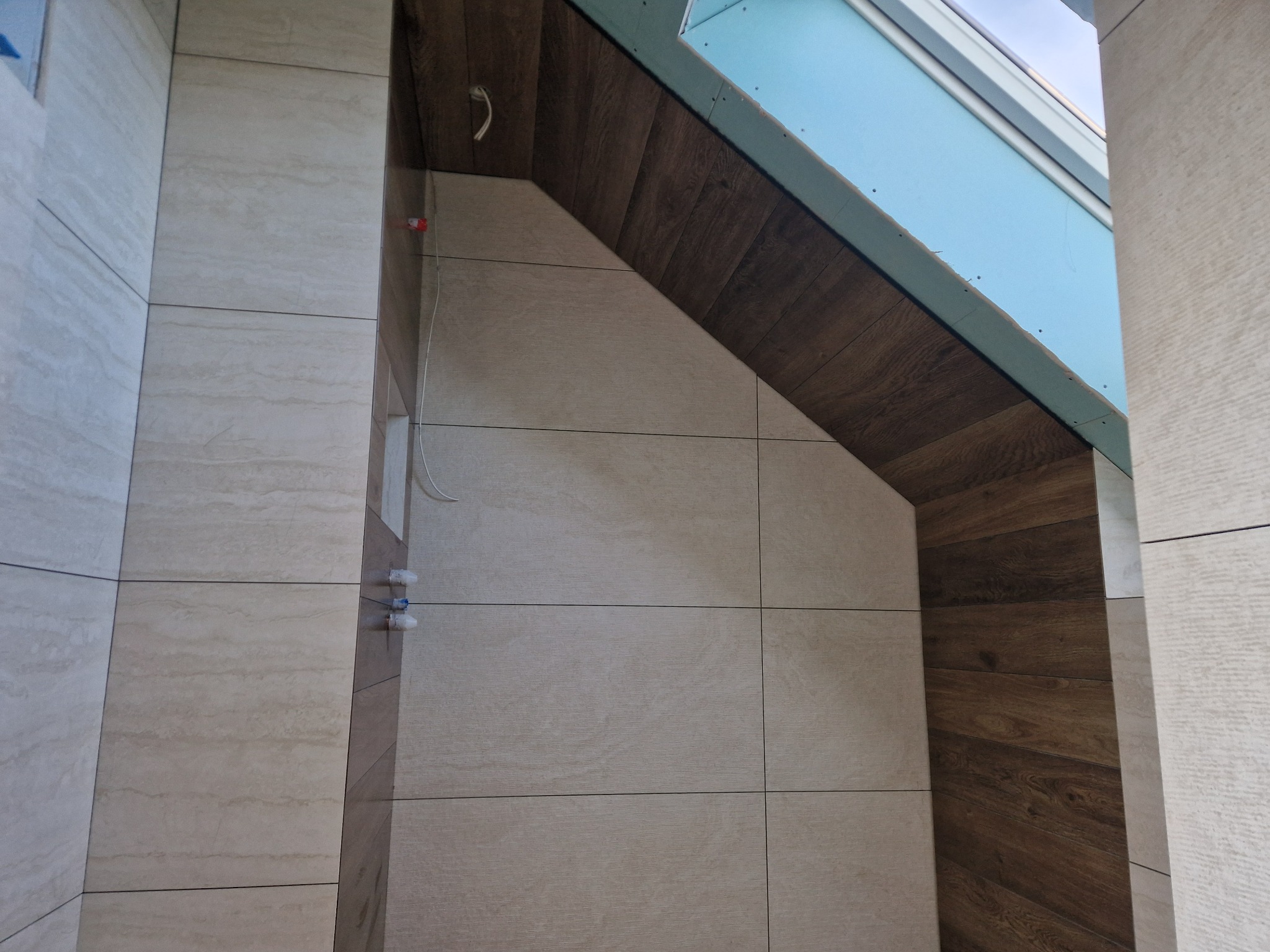 Wykończona łazienka na poddaszu z beżowymi płytkami imitującymi drewno na ścianach i ciemnobrązowymi płytkami na skosach. Widoczne instalacje wodne.