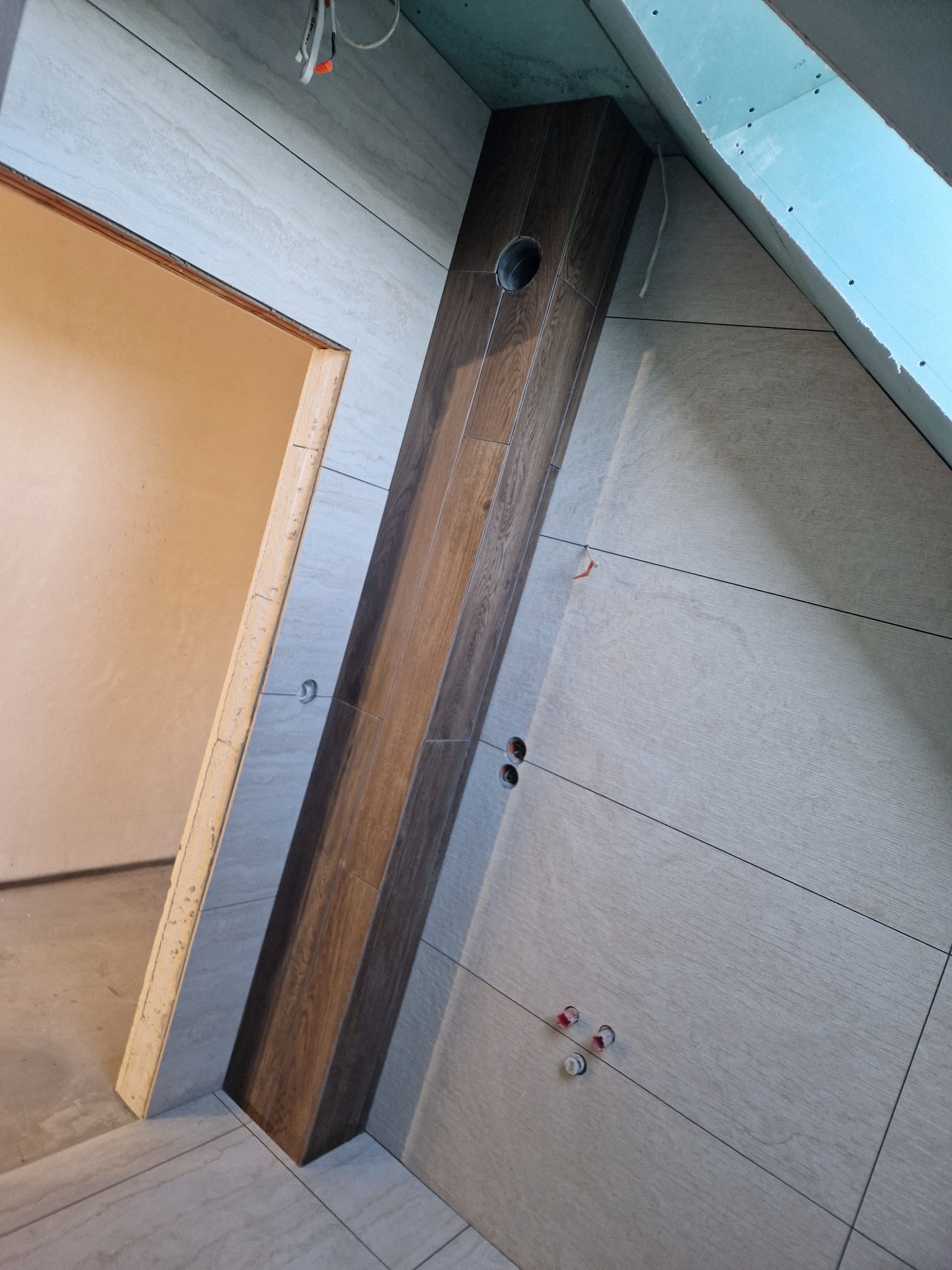 Wykończenie poddasza z płytami imitującymi drewno na ścianie i podłodze, pionowy element dekoracyjny z otworem, widoczne przewody i fragment okna dachowego.