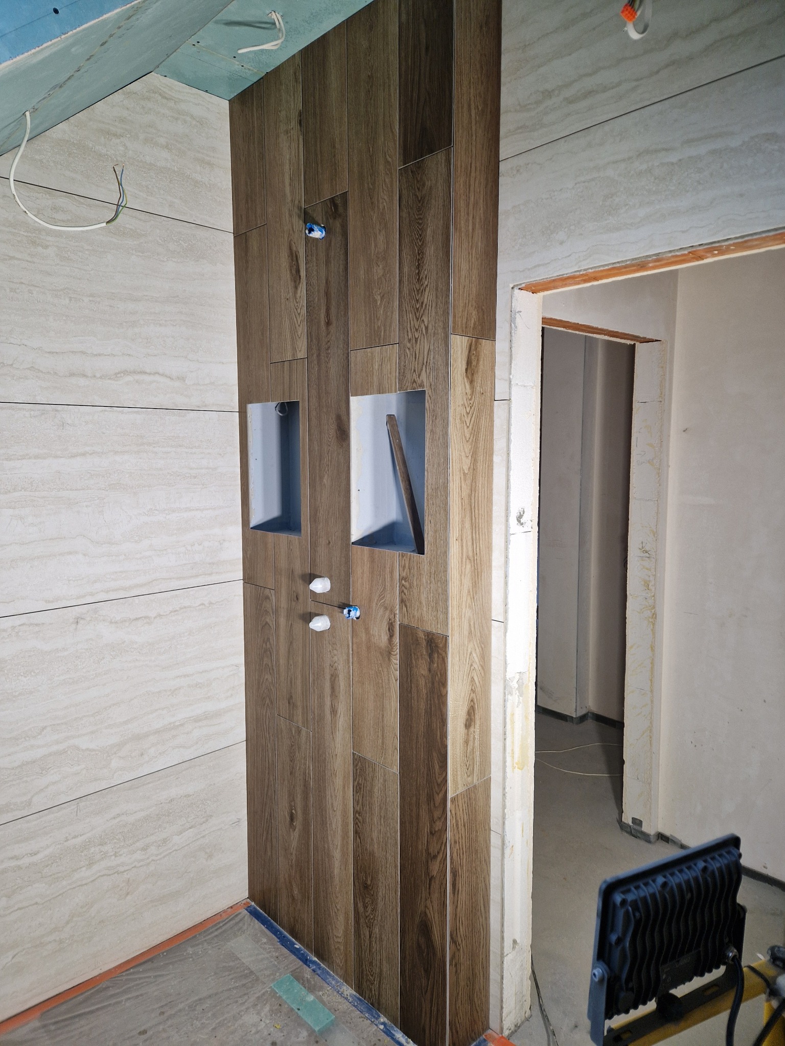 Ściana w łazience z drewnopodobnych płytek, wnęki z szarym wnętrzem, widoczne instalacje wodne. W tle fragment surowego pomieszczenia i oświetlenie budowlane.