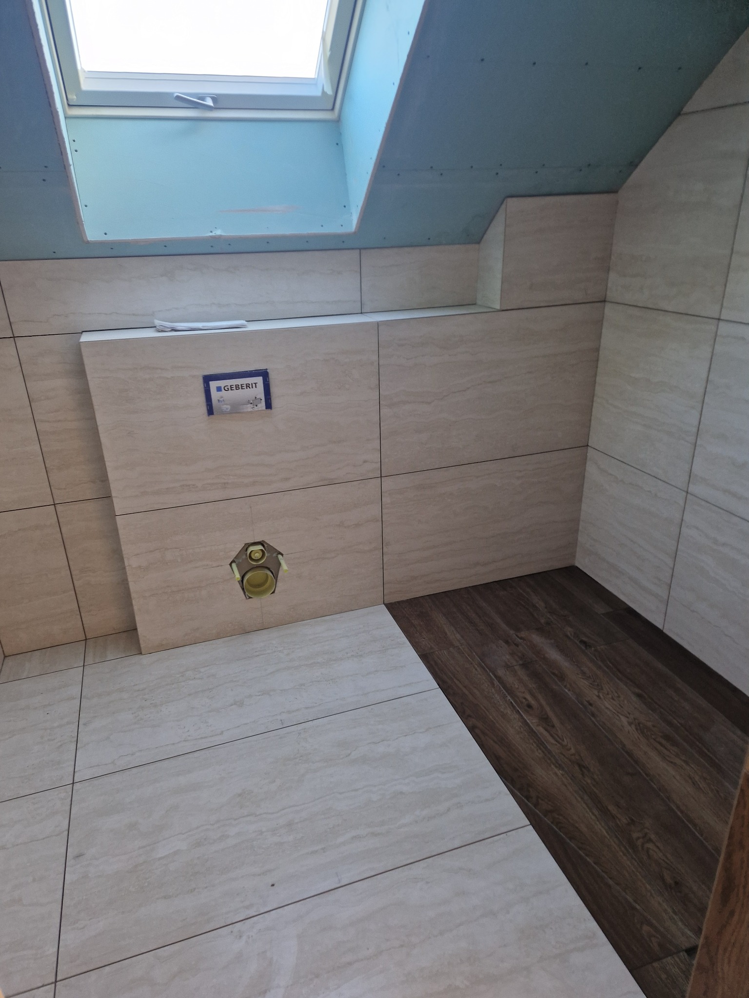 Wykończona łazienka z beżowymi płytkami na ścianach i podłodze, widoczna instalacja podtynkowa WC Geberit i fragment drewnopodobnej podłogi, okno dachowe.