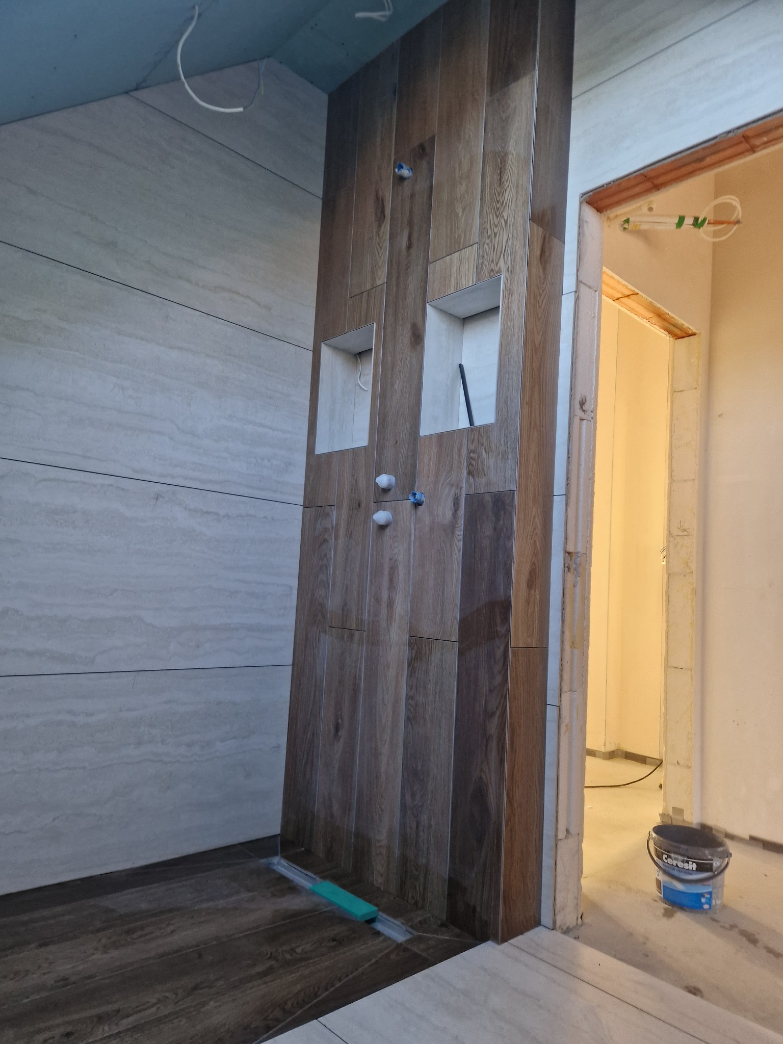 Wykończenie łazienki z drewnopodobną zabudową, wnękami i odpływem liniowym. Widoczne płytki imitujące drewno na ścianie i podłodze, oraz surowy stan wykończenia w sąsiednim pomieszczeniu.